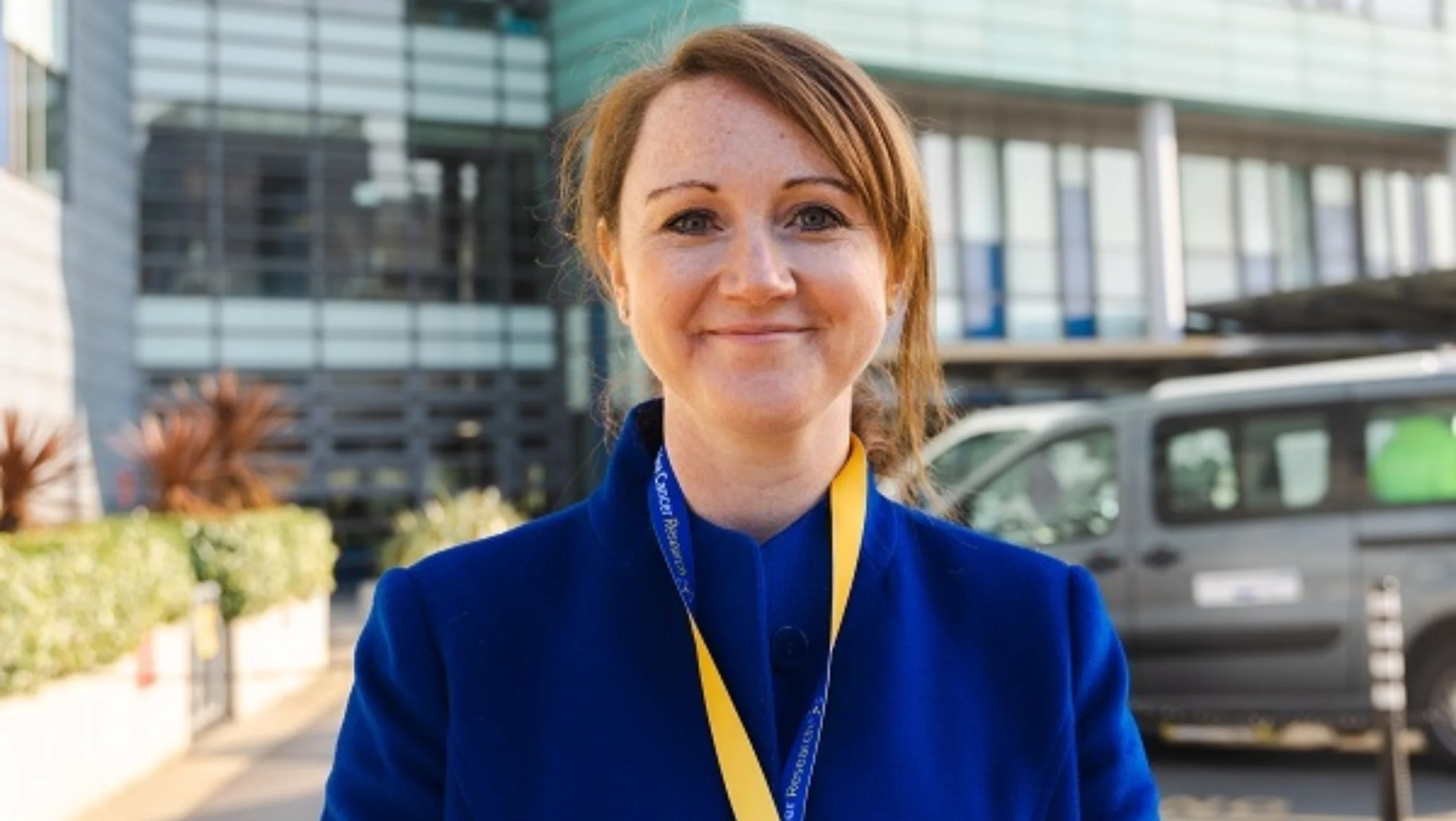 Jenny is wearing a blue coat and a yellow lanyard smiling at the camera outside a modern building.