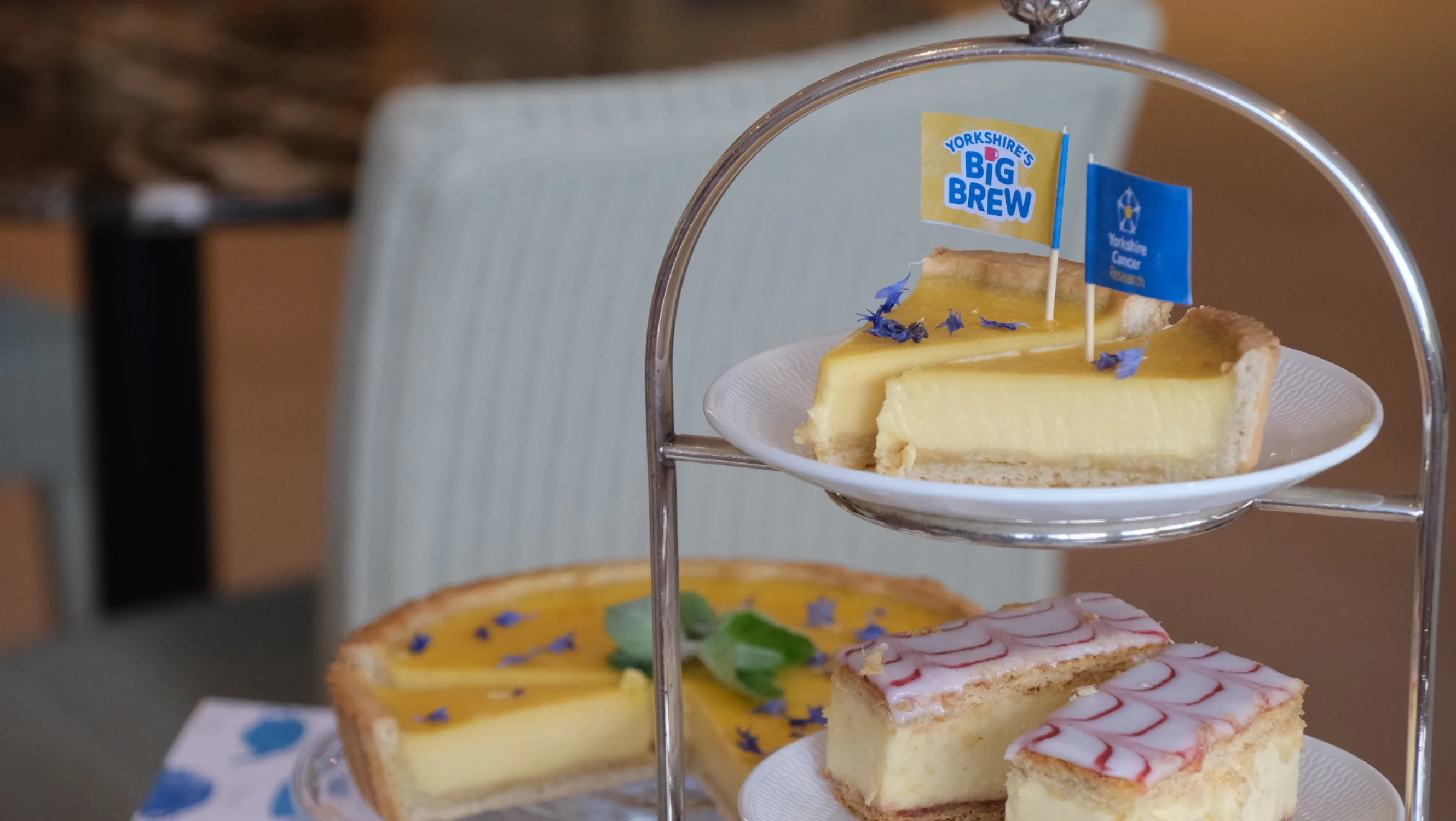 An afternoon tea stand with cakes and sweet treats, including slices of the Bettys-exclusive lemon tart. Two flags are on the top of the tart, including the Yorkshire's Big Brew and Yorkshire Cancer Research logos.