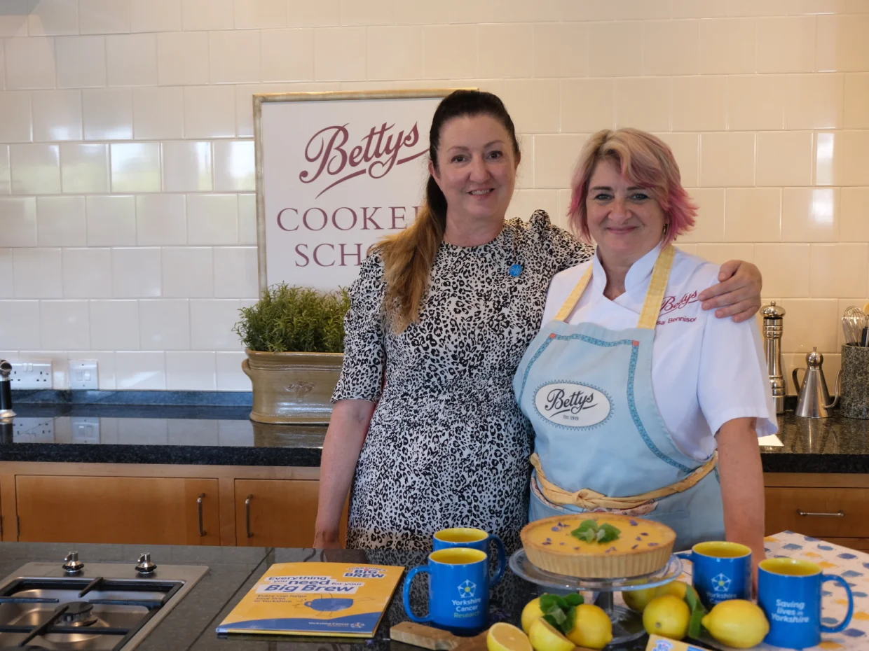 Ruth and Lisa from Bettys stand in Bettys Cookery School, with their arms around each other smiling at the camera. A lemon tart and Yorkshire Cancer Research merchandise is visible on the worktop in front.
