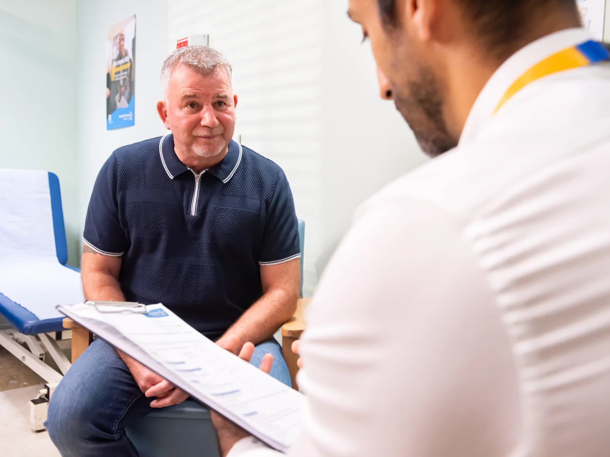 A patient sits in a medical setting while in conversation with a healthcare professional, who is wearing a Yorkshire Cancer Research lanyard and holding a clipboard.