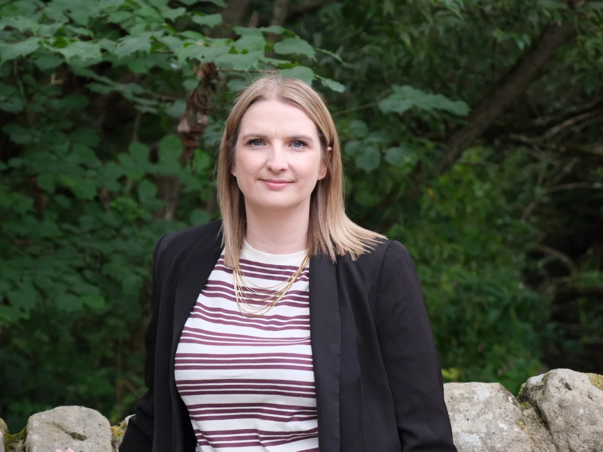 Beth Elias standing, facing the camera in a striped shirt, black blazer against a wall and trees